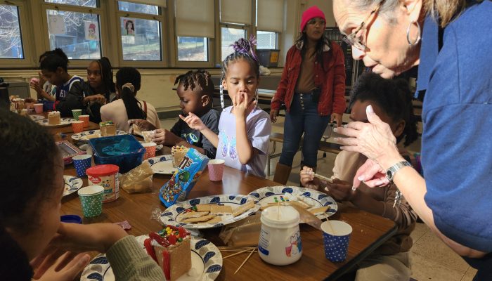 An adult looks over a table of supplies where children are assembling gingerbread houses